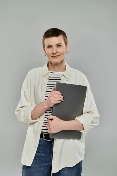 Une femme aux cheveux courts et au sourire détendu tient son ordinateur portable à la main, prête à travailler. — Photo de stock