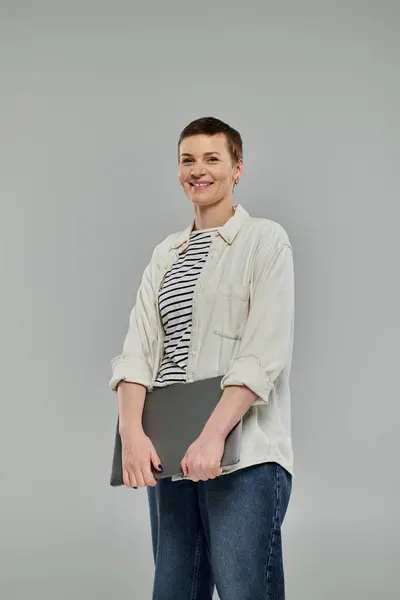 A woman with short hair and a white shirt smiles confidently while holding a laptop in front of a grey background. — Stock Photo