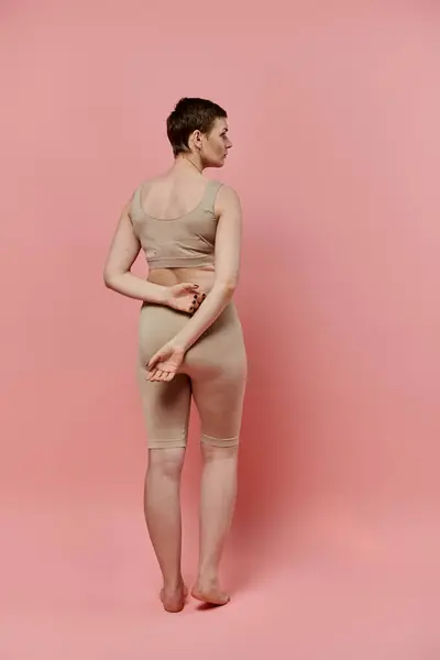 Una donna con i capelli corti, che indossa un reggiseno beige si trova di fronte alla telecamera in uno studio rosa. — Foto stock