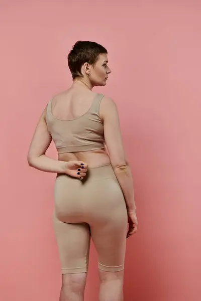 A woman with short hair, wearing a beige sports bra and shorts, stands facing away from the camera in her bra. — Stock Photo