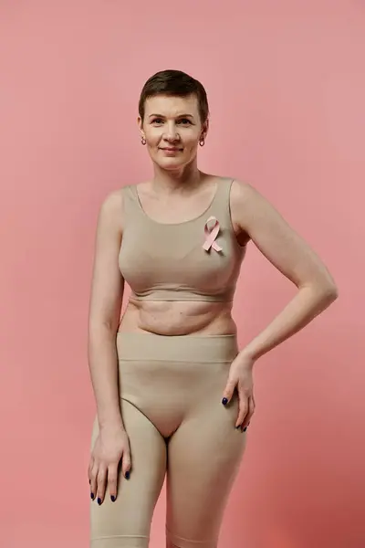 Donna dai capelli corti in reggiseno beige e pantaloncini posa con fiducia sullo sfondo rosa, con un nastro rosa appuntato al petto — Foto stock