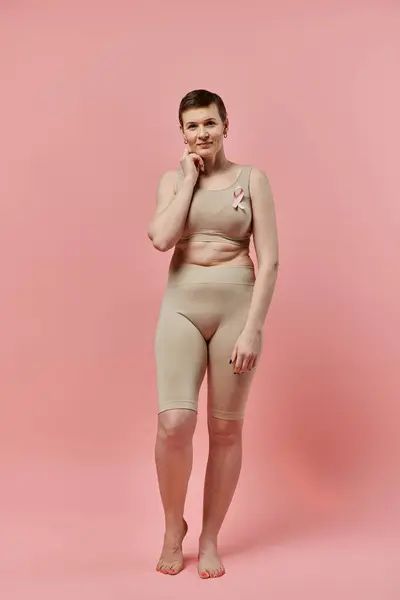 Una donna con i capelli corti si trova in un reggiseno beige e pantaloncini, un nastro rosa appuntato al petto, su uno sfondo rosa. — Foto stock