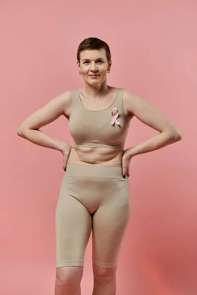 A woman with short hair stands confidently in a bra and shorts, a pink ribbon pinned to her chest. — Stock Photo