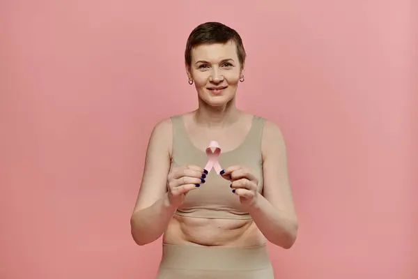 Una mujer con el pelo corto, usando un sujetador y sosteniendo una cinta rosa, sonríe a la cámara. — Stock Photo