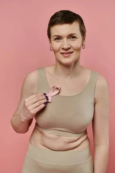 A woman with short hair, wearing a bra, holds a pink ribbon to her chest, a symbol of breast cancer awareness and support. — Stock Photo