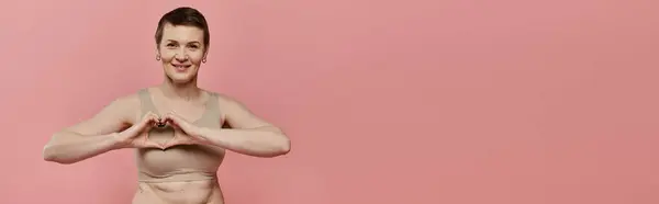 A woman with short hair wearing a bra with a pink ribbon makes a heart shape with her hands in front of a pink background. — Stock Photo