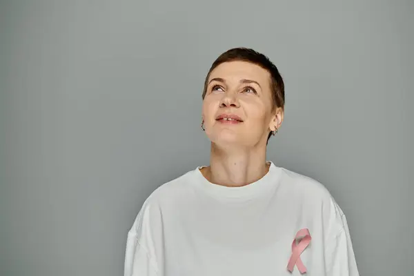 Eine Frau mit kurzen Haaren blickt nach oben, ihr weißes Hemd ziert eine rosafarbene Schleife, ein Symbol für Brustkrebs-Aufklärung und -Prävention.. — Stockfoto