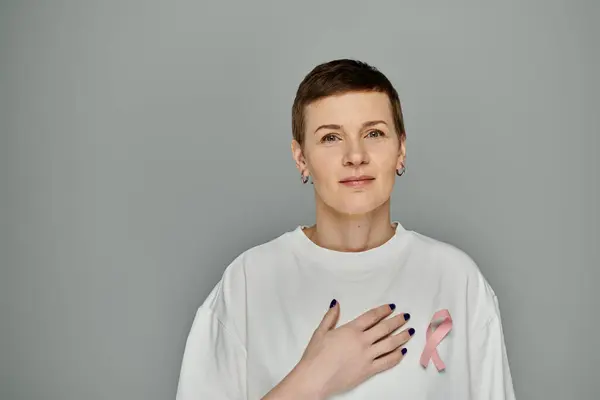 Uma mulher com cabelo curto e uma fita rosa em sua camisa, de pé contra um fundo cinza. — Fotografia de Stock