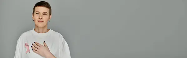A woman with short hair, wearing casual attire and a pink ribbon, stands against a grey background, advocating for breast cancer awareness. — Stock Photo