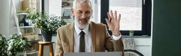 Un maturo, barbuto gay uomo in un abito sorride e onde mentre insegna online utilizzando un computer portatile. — Foto stock