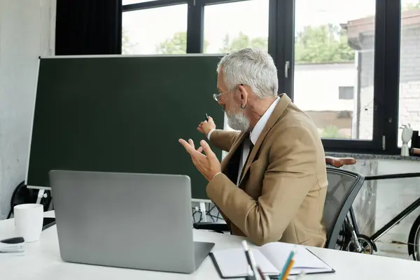 A middle-aged man with a beard teaches online, gesturing towards his laptop with a blackboard behind him, lgbtq teacher — Stock Photo