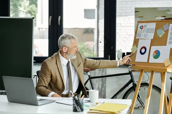 A mature gay teacher with a beard explains charts to his online students, sitting at a desk with a laptop. — Stock Photo
