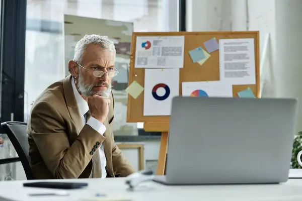 A bearded man sits at a desk, thoughtfully looking at a laptop screen while teaching online. — Stock Photo