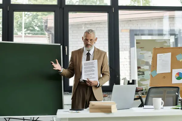 Un uomo di mezza età con la barba, vestito con un abito, istruisce gli studenti attraverso un computer portatile in un ambiente di classe, insegnante lgbtq — Foto stock