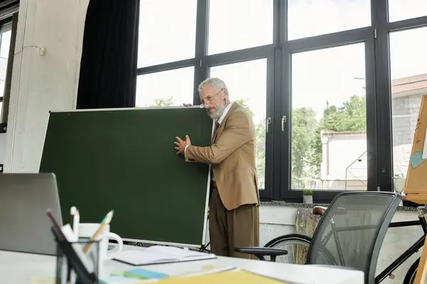 Зріла, бородата вчителька ЛГБТ в коричневому костюмі готується до онлайн уроку в сучасному, яскравому класі. — стокове фото
