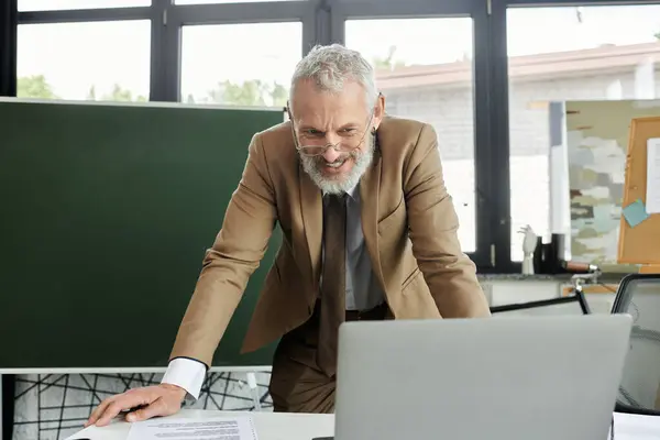 A mature man with a beard, wearing a suit, smiles as he teaches an online class using a laptop in a classroom, lgbtq teacher — Stock Photo