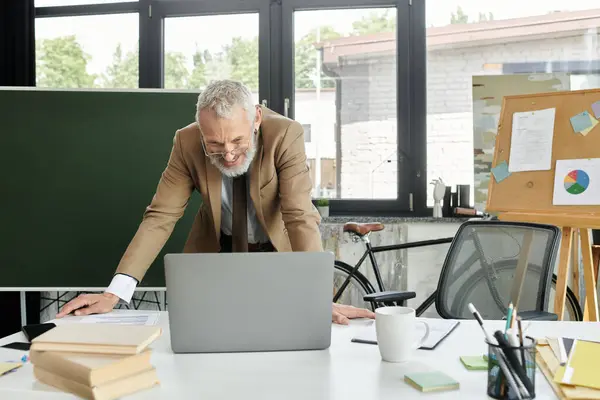 Un insegnante di mezza età con la barba sorride mentre insegna online, appoggiato a un computer portatile su un tavolo pieno di libri e forniture per l'ufficio. — Foto stock
