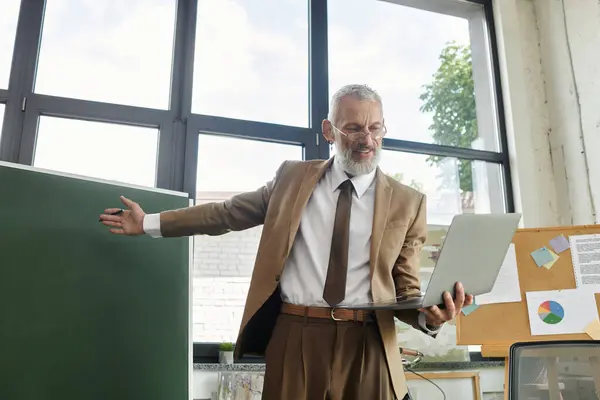 A bearded, mature teacher in a classroom setting teaches students online through his laptop. — Stock Photo