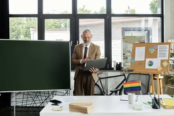 A mature LGBTQ teacher with a beard uses a laptop to teach online from a classroom with a blackboard, a bicycle, and a rainbow flag. — Stock Photo