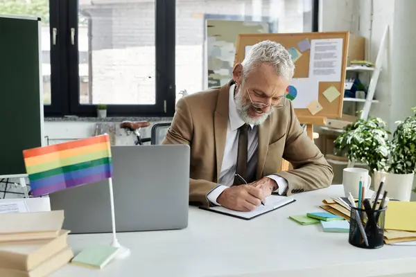 A mature LGBTQ+ teacher with a beard writes in a notebook at his desk, a rainbow flag in the foreground. — Stock Photo
