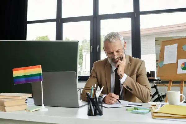 A middle-aged man with a beard sits at a desk with a laptop and a rainbow flag, thoughtfully writing in a notebook. — Stock Photo
