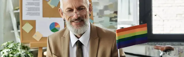 A mature man with a beard smiles while teaching online, a rainbow flag prominently displayed behind him. — Stock Photo