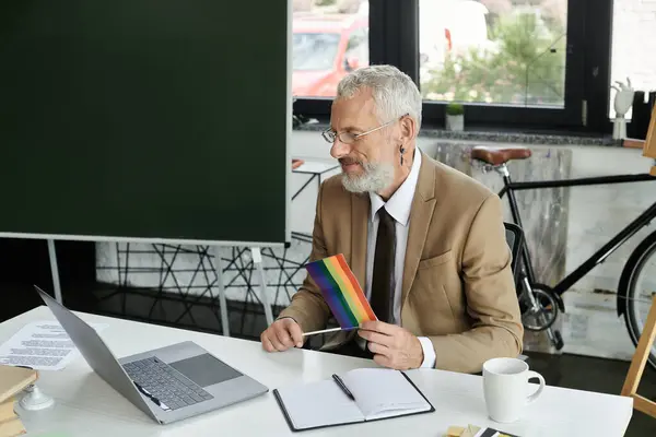 A mature teacher with a beard sits at a desk in a classroom, teaching online via laptop, holding a rainbow flag. — Stock Photo