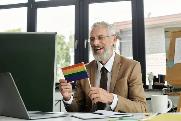 A mature LGBTQ teacher with a beard, smiles as he holds up a pride flag during an online class. — Stock Photo