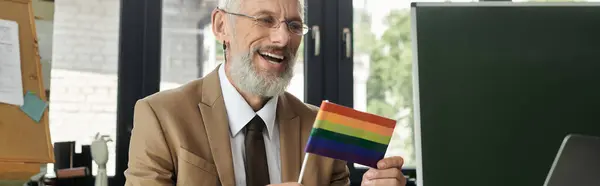 A smiling, bearded, middle-aged gay teacher holds a rainbow flag while teaching online in a classroom. — Stock Photo