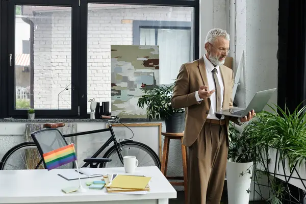 A middle-aged man with a beard, wearing a suit and tie, uses a laptop to teach an online class. A rainbow flag adorns the desk. — Stock Photo