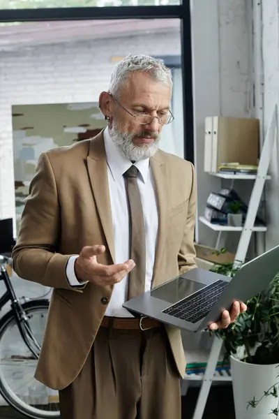 A middle-aged man with a beard, wearing a suit, teaches online through a laptop in a bright, modern office. — Stock Photo