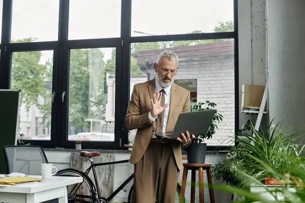 Ein bärtiger Mann in Anzug und Krawatte unterrichtet online per Laptop, während er in einem hellen, modernen Büro mit Pflanzen steht.. — Stockfoto