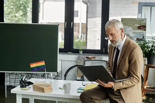 A middle-aged man with a beard and glasses teaches online in a classroom, a rainbow flag displayed prominently on a desk. — Stock Photo