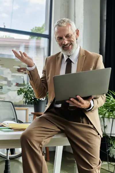 A mature man with a beard sits at a desk, holding a laptop while smiling and gesturing with his hand. — Stock Photo