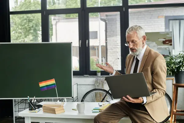 Un profesor LGBTQ maduro con barba enseña en línea a través de un portátil en un aula moderna. - foto de stock