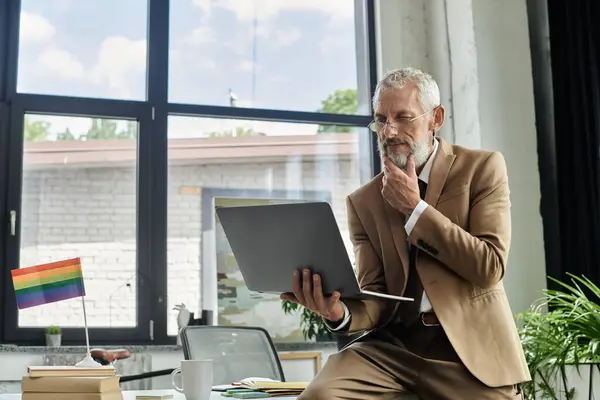 Bearded man in front of a window at a desk, engrossed in an online class on his laptop, with a pride flag nearby — Stock Photo