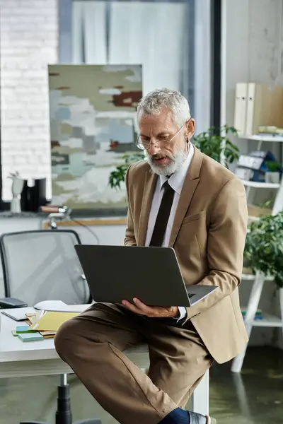 Un uomo maturo con la barba si siede su una scrivania, guardando attentamente il suo computer portatile, mentre insegna online. — Foto stock