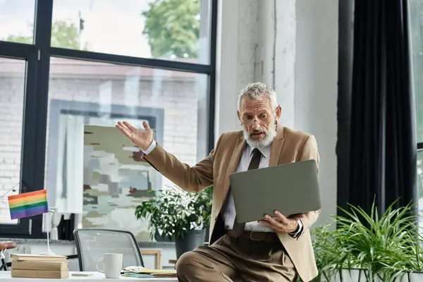 Un insegnante LGBTQ maturo, indossando un completo, spiega appassionatamente una lezione mentre tiene un computer portatile in un ambiente di ufficio.. — Foto stock