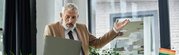 A mature, bearded teacher in a suit passionately teaches a lesson online, his hand gesturing toward the laptop screen. — Stock Photo