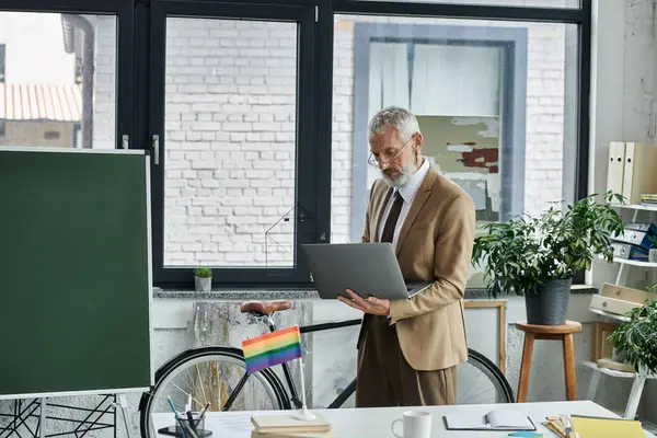 A mature, bearded man in a suit stands in a bright room, holding a laptop and preparing to teach online. — Stock Photo