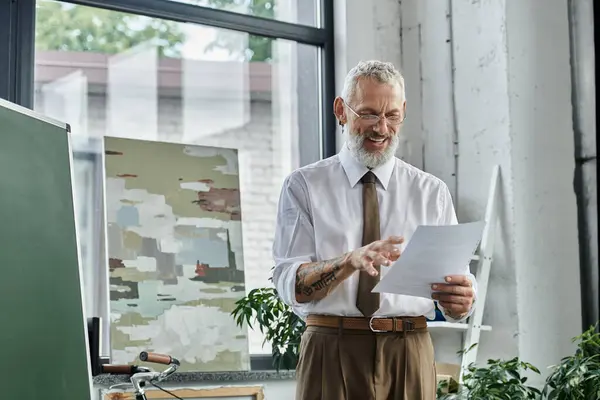 A mature, bearded teacher in a white shirt and tie smiles as he reads a paper, likely for an online class. — Stock Photo