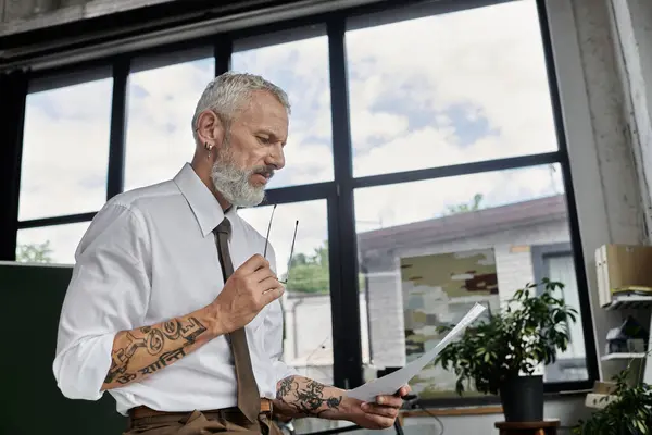 A mature man with a beard and tattoos prepares to teach an online class, reviewing notes while holding a pair of glasses in his hand. — Stock Photo
