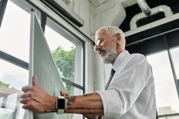 A mature, bearded teacher in a white shirt writes equations on a whiteboard, teaching online. — Stock Photo