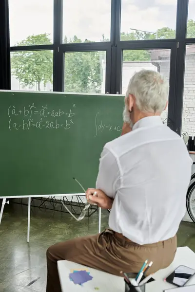 A mature LGBTQ teacher with a beard sits by a chalkboard during an online lecture. — Stock Photo
