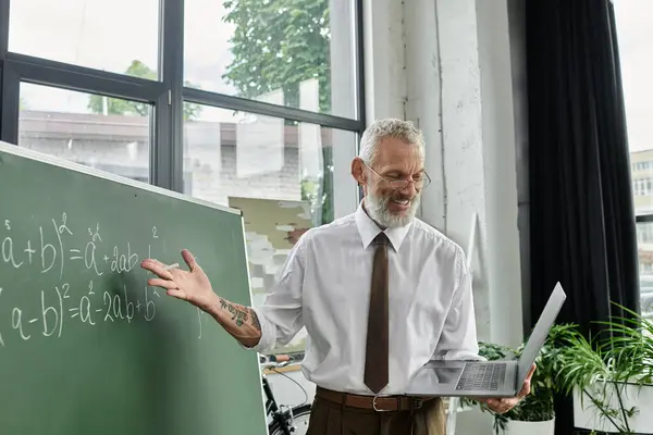 A middle-aged man with a beard teaches mathematics online, using a laptop and a chalkboard. — Stock Photo