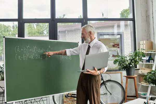 Bearded man in white shirt and brown pants teaches math online with laptop in front of blackboard with equations — Stock Photo