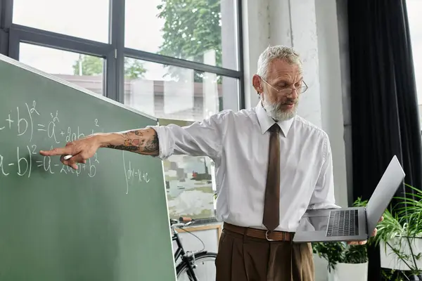 A mature, bearded teacher passionately explains a math equation on a chalkboard while using a laptop for online instruction. — Stock Photo