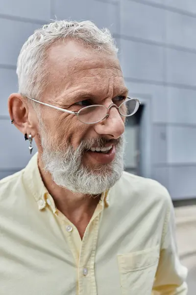 A mature gay man with a white beard and glasses smiles while standing outdoors near a modern building. — Stock Photo