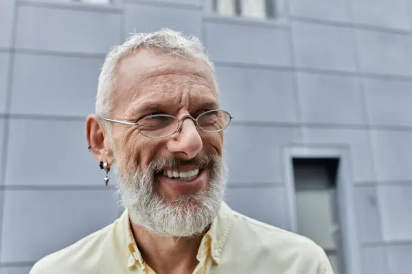 Зрілий гей чоловік з білою бородою, одягнений в окуляри, посміхається, стоячи перед сучасною будівлею. — стокове фото