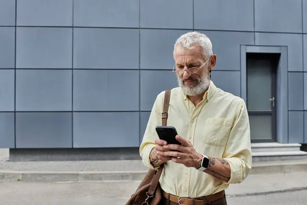 A mature man with a beard and glasses checks his phone while standing in front of a modern building. — Stock Photo